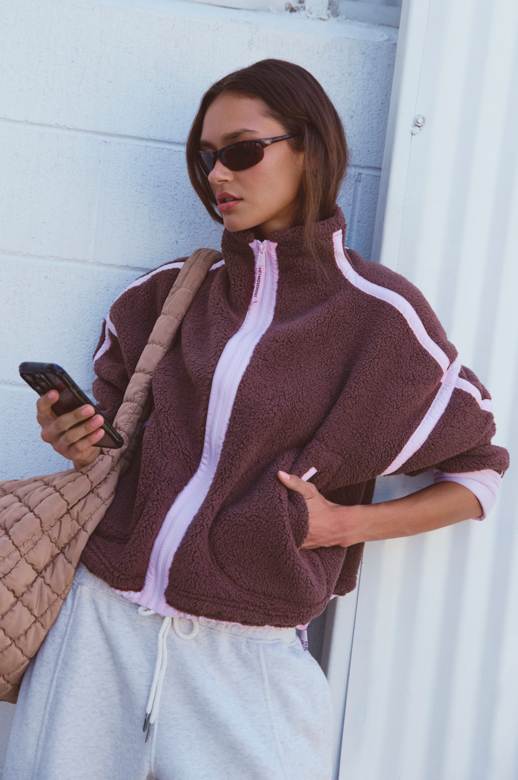 A woman wearing a brown sherpa fleece zip-up jacket with light pink trim and light gray sweatpants, holding a phone and carrying a quilted tan shoulder bag against a white paneled wall.