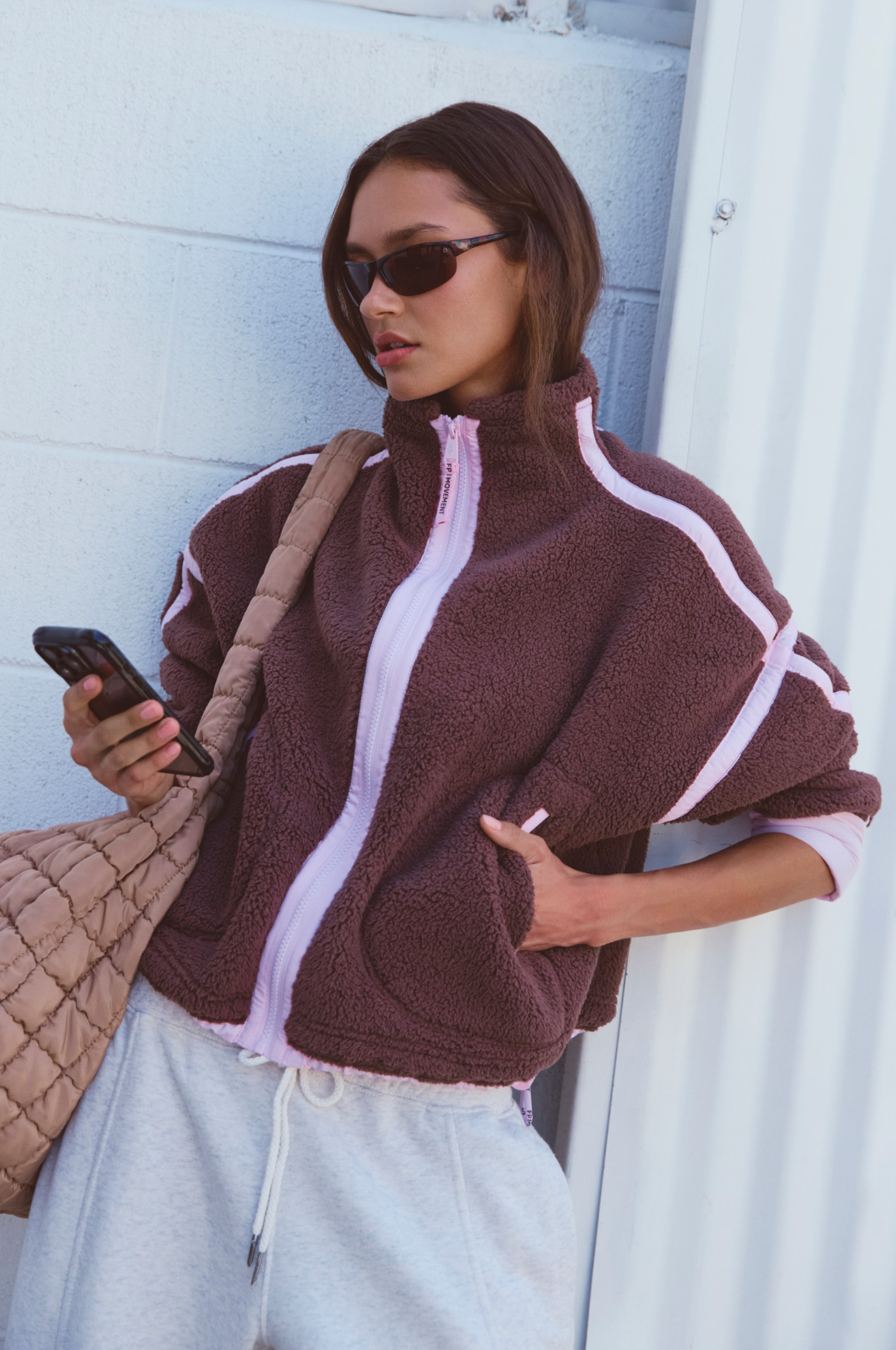 A woman wearing a brown sherpa fleece zip-up jacket with light pink trim and light gray sweatpants, holding a phone and carrying a quilted tan shoulder bag against a white paneled wall.