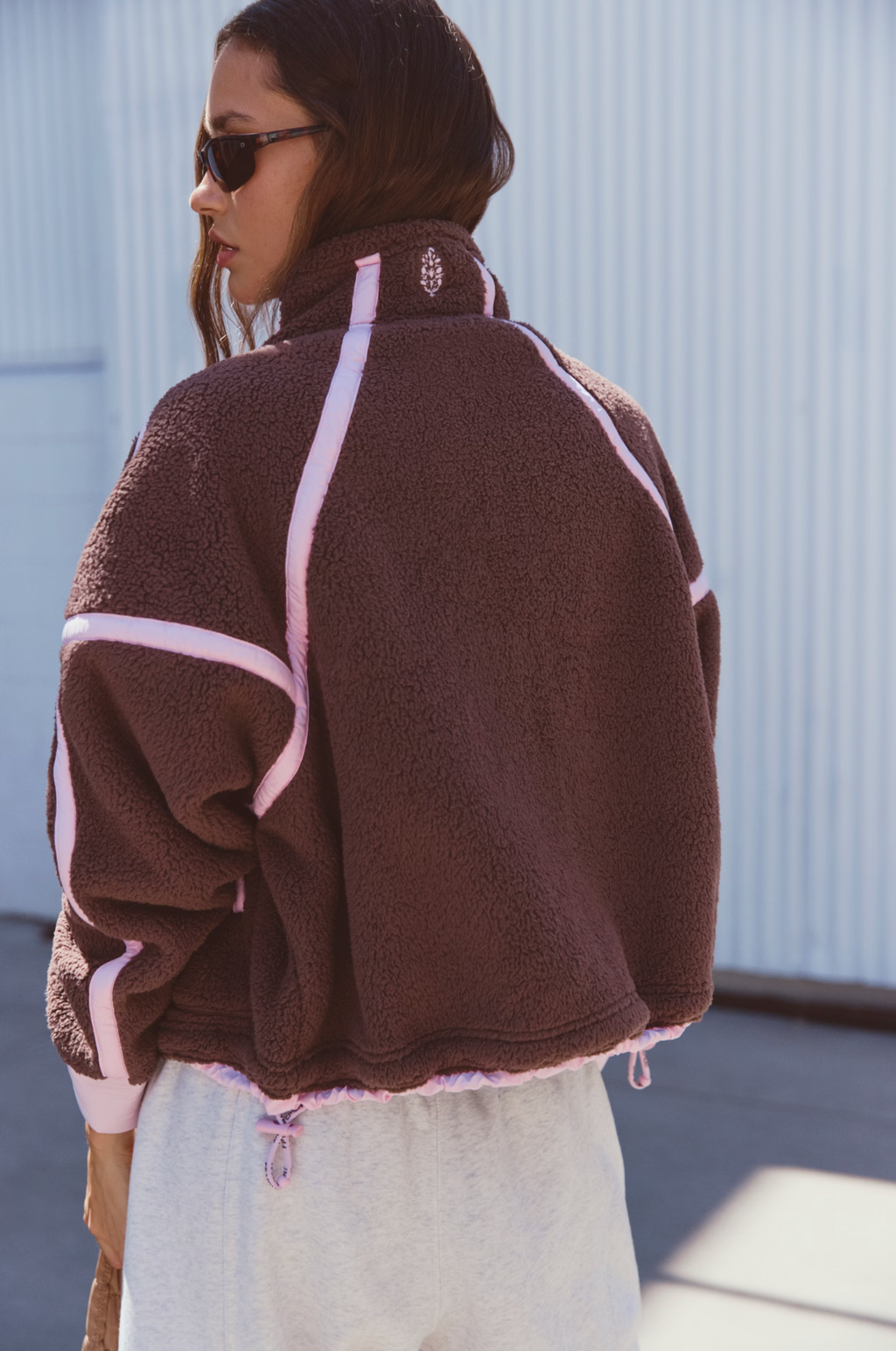 A woman wearing a brown sherpa fleece jacket with light pink contrast stitching, shown from the back in an outdoor setting against a light blue corrugated metal wall.