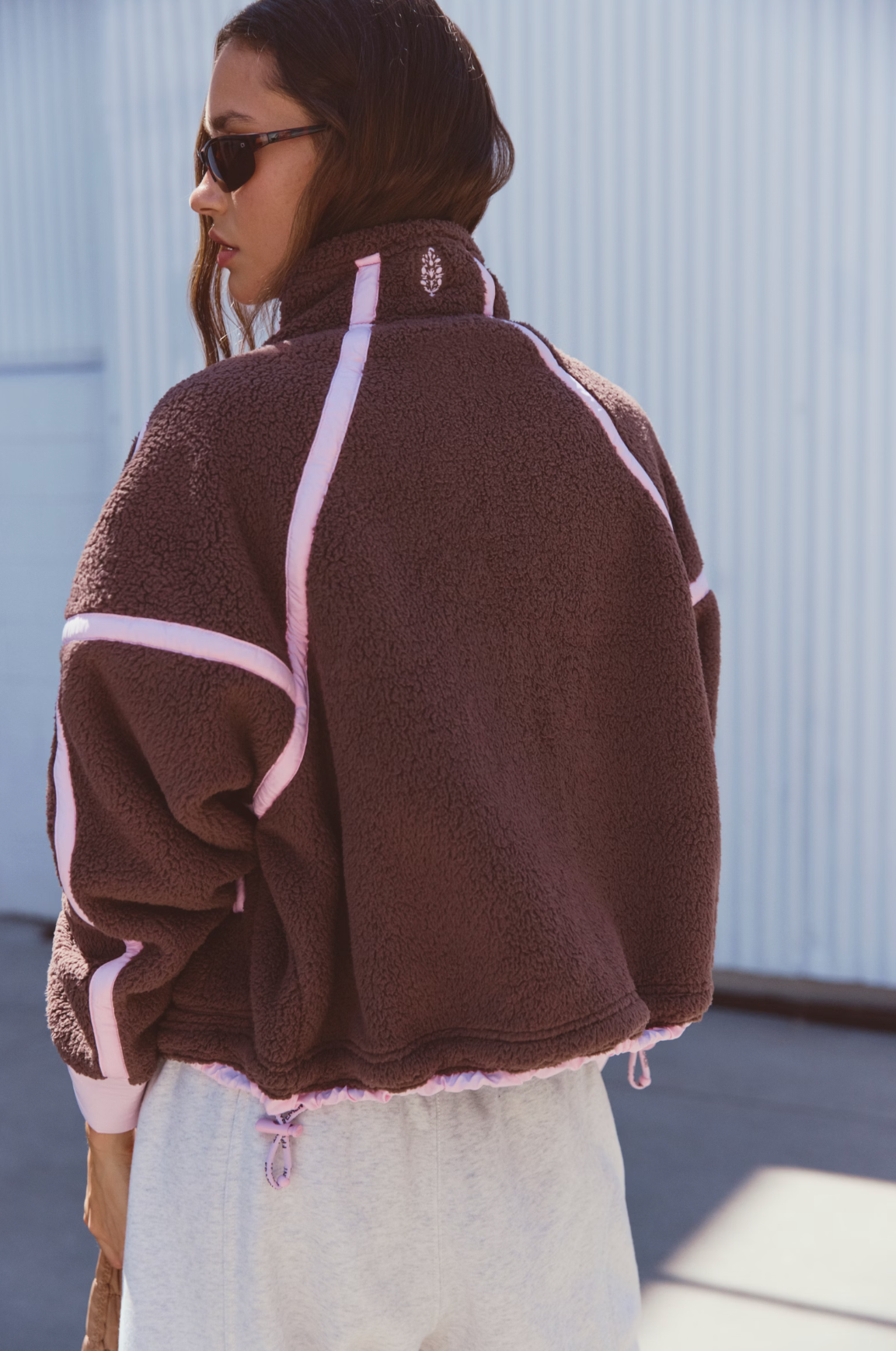 A woman wearing a brown sherpa fleece jacket with light pink contrast stitching, shown from the back in an outdoor setting against a light blue corrugated metal wall.