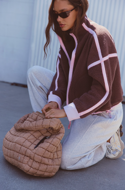 A woman kneels on a concrete surface wearing a brown fleece zip-up jacket with light pink trim, light gray sweatpants, and white sneakers, while holding a large quilted tan shoulder bag.
