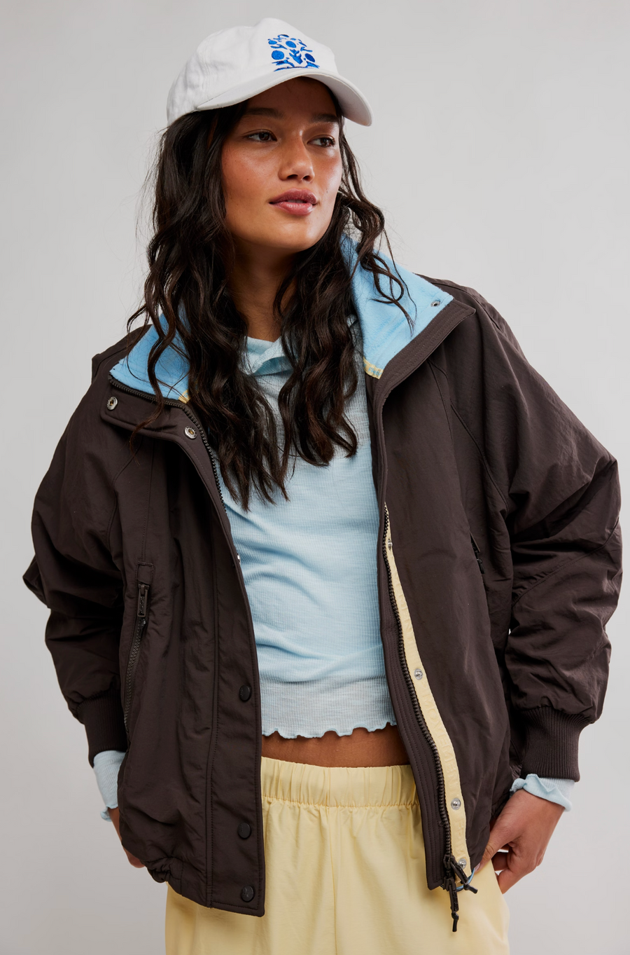 A model wearing a dark brown lightweight jacket with a light blue inner collar, styled over a light blue crop top and cream-colored pants, paired with a white cap featuring a blue graphic.