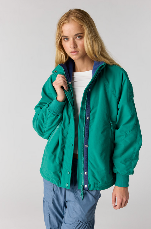 A young woman modeling a bright green lightweight jacket with a dark blue inner lining and snap-button front, worn over a white top and blue pants, against a light gray studio background.