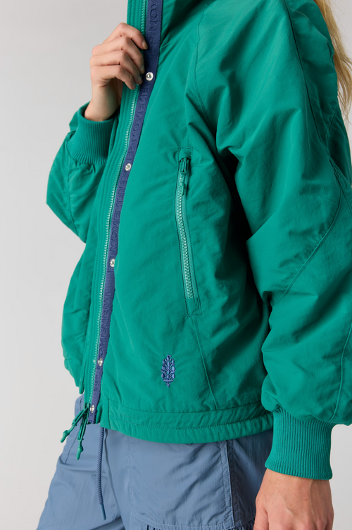 A close-up of a person wearing a teal green bomber jacket with a purple zipper and a small embroidered logo on the front, paired with blue cargo pants against a light gray studio background.