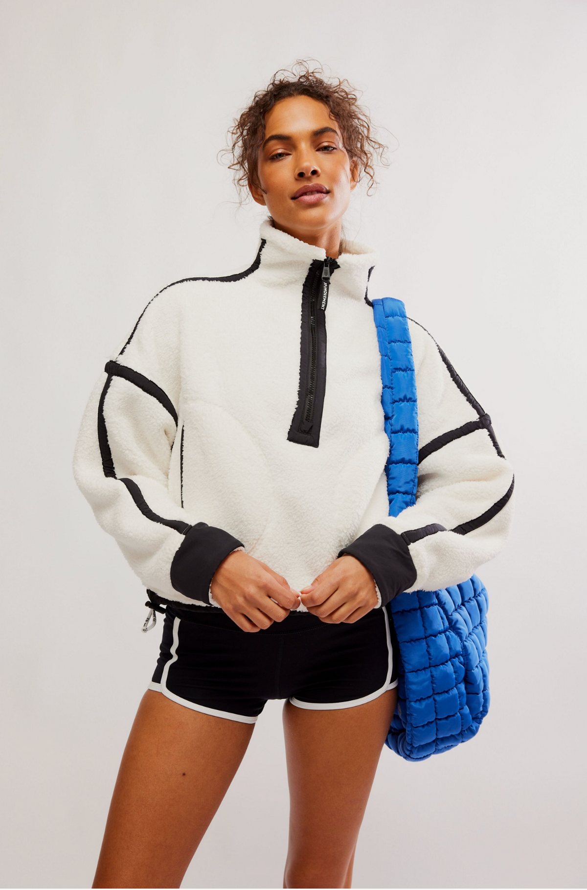 A model wearing a white fleece half-zip pullover with black trim, black athletic shorts, and carrying a bright blue quilted shoulder bag against a plain light gray studio background.