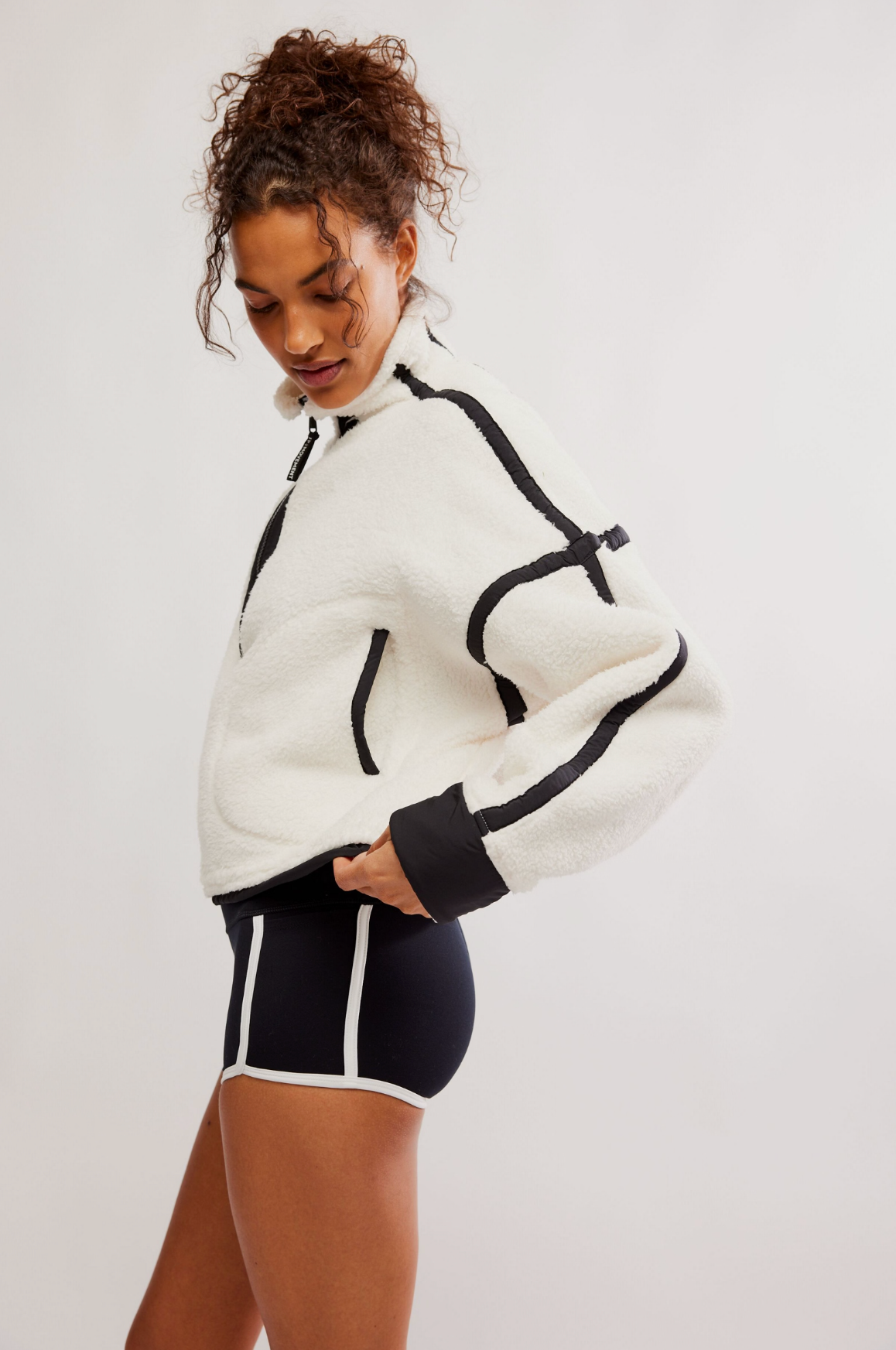 A woman wearing a white fuzzy cropped zip-up jacket with black contrast piping and black athletic shorts with white side stripes, posed against a light gray studio background.