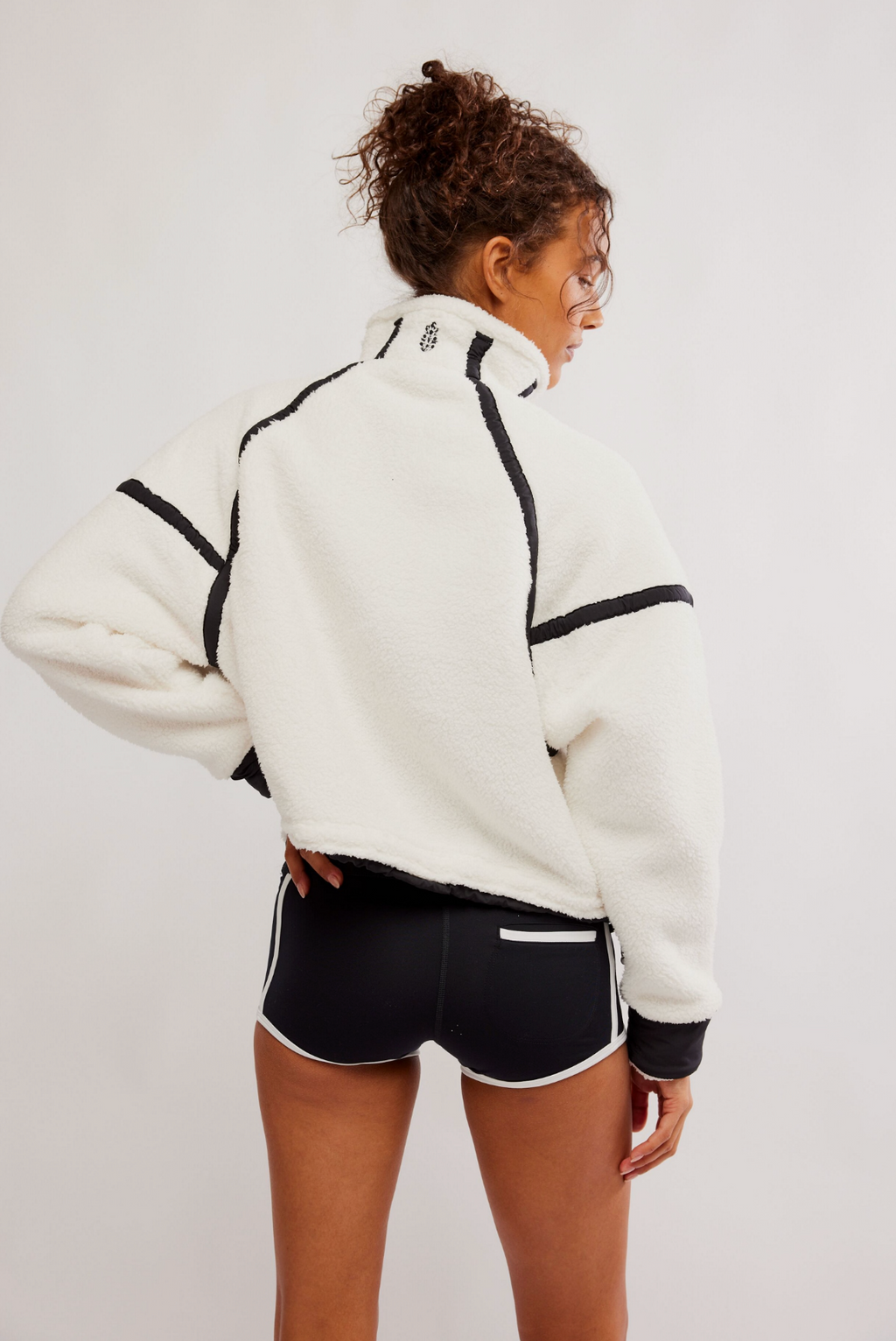 A woman shown from the back wearing a white sherpa fleece cropped jacket with black contrast piping and black athletic shorts with white side stripes, against a plain light gray studio background.