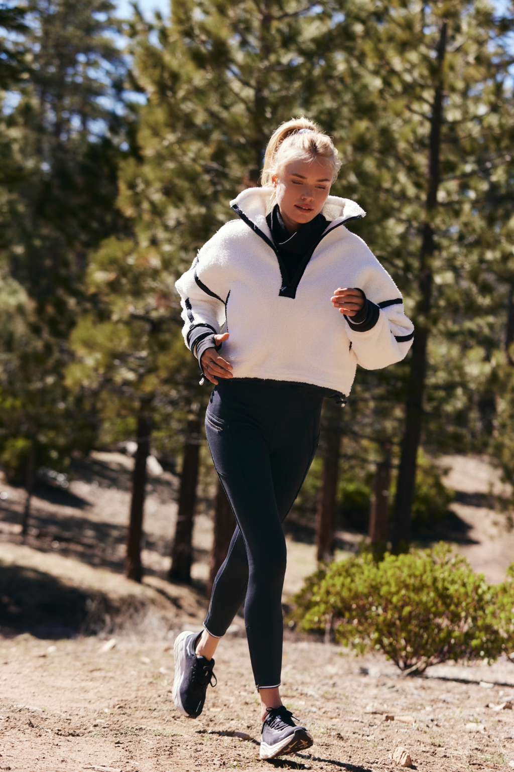 A woman in a white cropped zip-up hoodie with black trim and black leggings stretches her leg outdoors in a wooded area. She is wearing black athletic shoes.