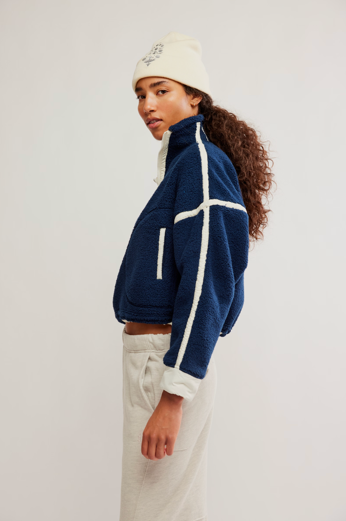 A woman wearing a navy blue cropped fleece jacket with white contrast stitching, paired with cream sweatpants and a cream beanie, photographed against a plain light gray studio background.