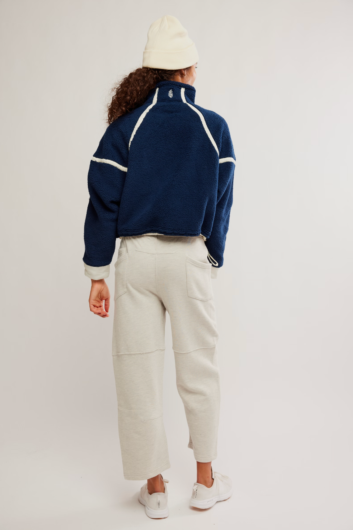 A model shown from the back wearing a navy blue cropped fleece jacket with white contrast stitching, cream-colored cropped pants, a cream beanie, and white sneakers, set against a plain light gray studio background.