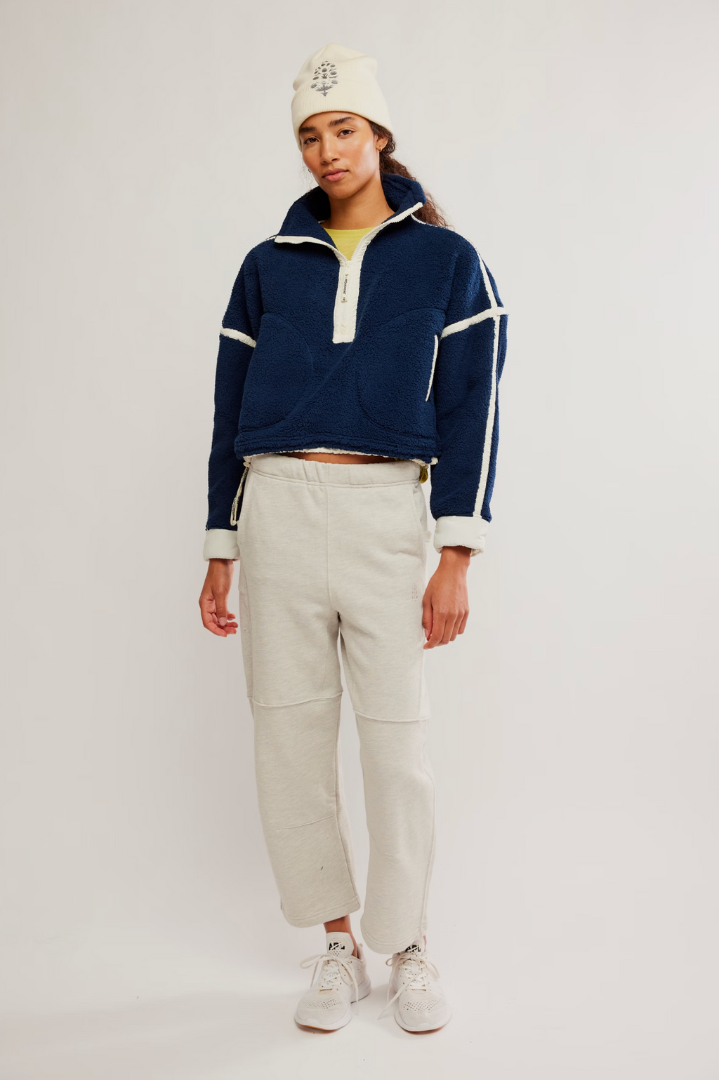 A model stands against a light gray studio background wearing a navy cropped quarter-zip fleece pullover with white stripe details, off-white sweatpants, a cream knit beanie, and white sneakers.