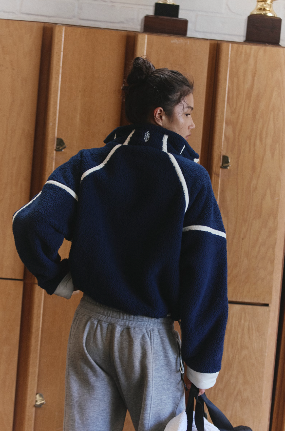 A person shown from the back wearing a navy blue fleece zip-up jacket with white stripe accents and gray sweatpants, standing in front of wooden lockers.