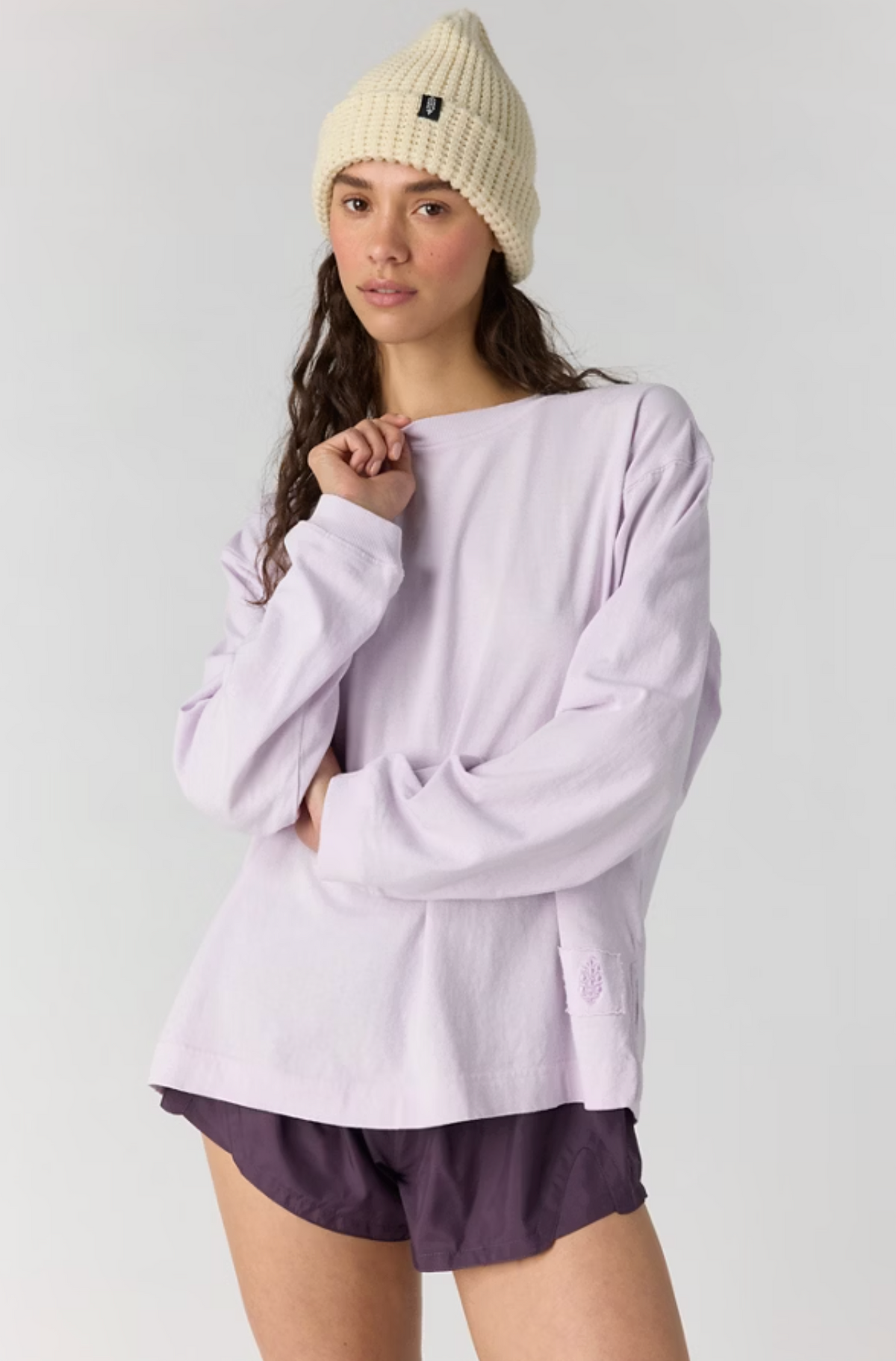 A model wearing a light lavender long-sleeve crewneck shirt, dark purple athletic shorts, and a cream-colored knit beanie hat, posed against a solid light gray background.