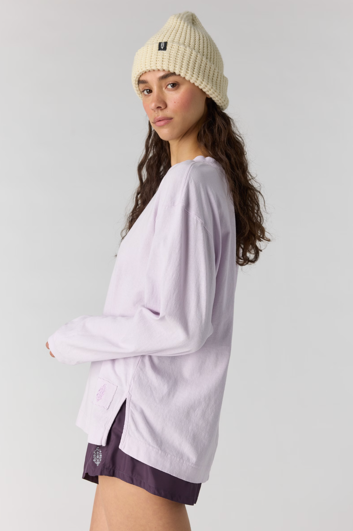 A woman wearing a light lavender long-sleeve shirt, dark purple shorts, and a cream-colored knit beanie hat, photographed against a solid light gray studio background.