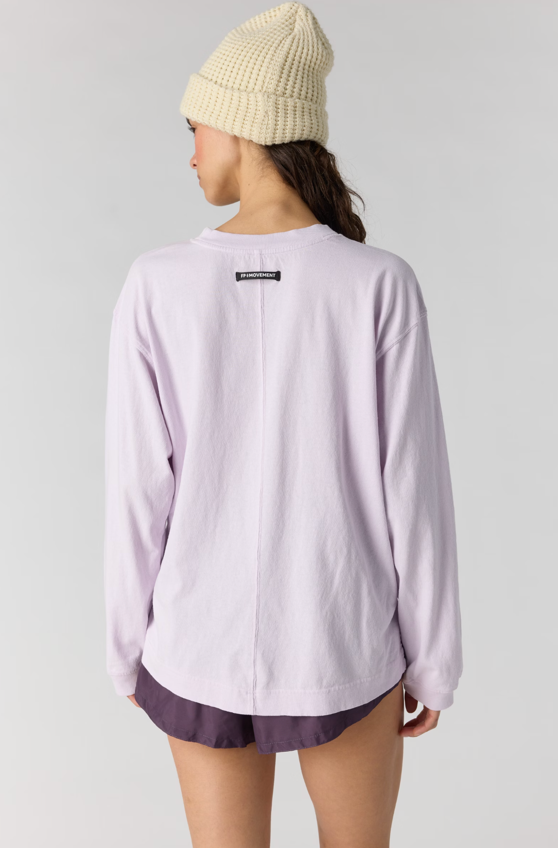 A model shown from the back wearing a light lavender long-sleeve crewneck sweatshirt, dark purple shorts, and a cream-colored chunky knit beanie against a light gray studio background.