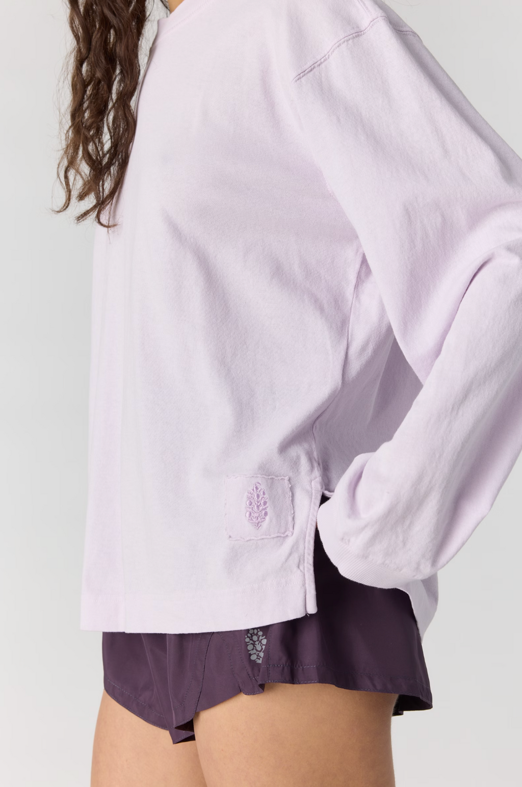 A close-up of a person wearing a light lavender long-sleeve top and dark purple shorts against a plain light gray background.