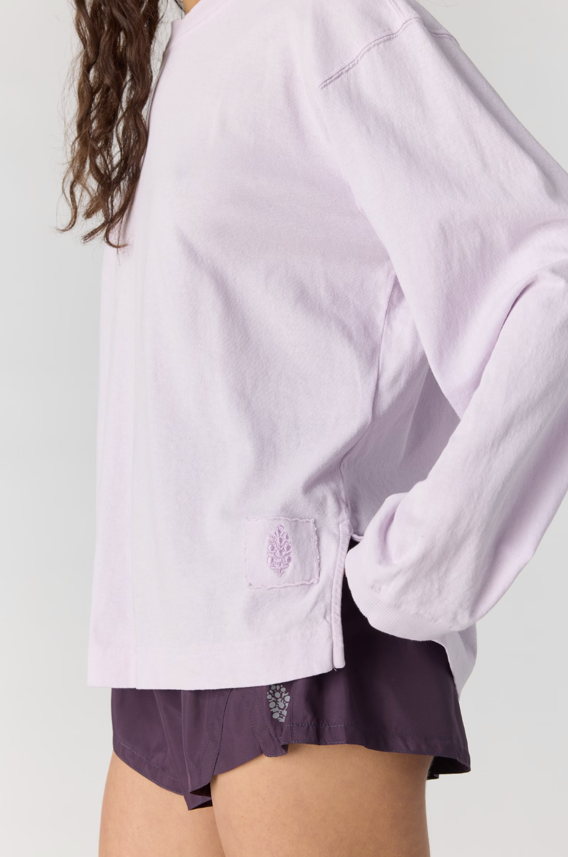 A close-up of a person wearing a light lavender long-sleeve top and dark purple shorts against a plain light gray background.
