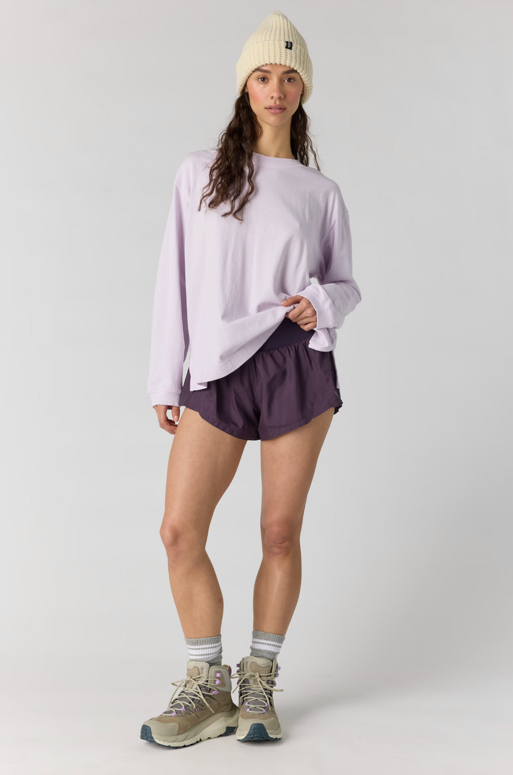 A model stands against a light gray studio background wearing a light purple oversized long-sleeve shirt, dark purple athletic shorts, a cream beanie, gray socks, and hiking shoes.