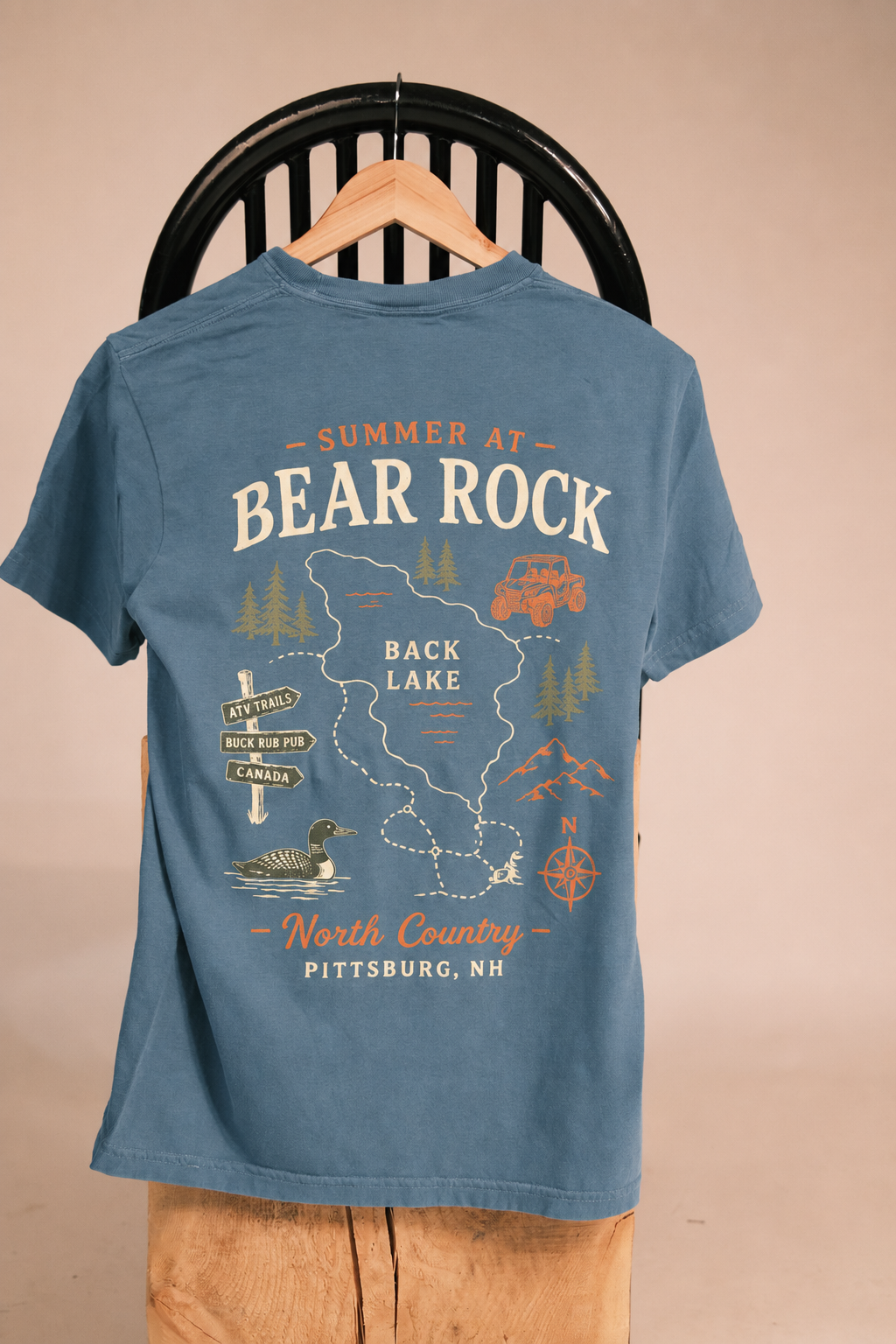 A blue graphic t-shirt displayed on a wooden hanger against a beige wall, with a black chair back visible behind it. The back of the shirt features a large vintage-style map graphic of Back Lake in Pittsburg, NH, with illustrations of a car, duck, deer, and compass rose.