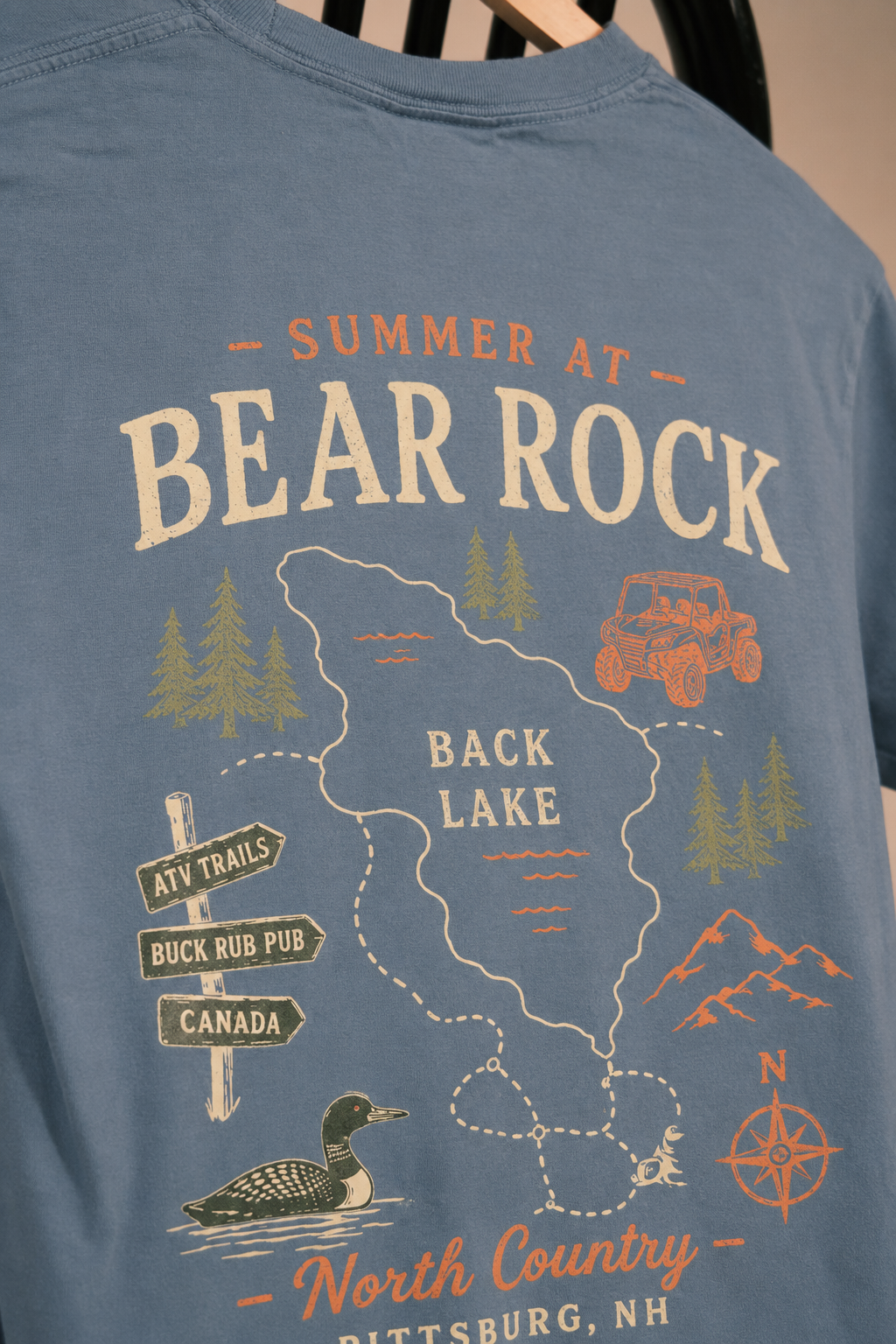 A close-up of a blue graphic t-shirt featuring a vintage-style outdoor map design with the text 'SUMMER AT BEAR ROCK' and 'BACK LAKE', along with illustrations of a car, trees, mountains, a duck, and directional signs.