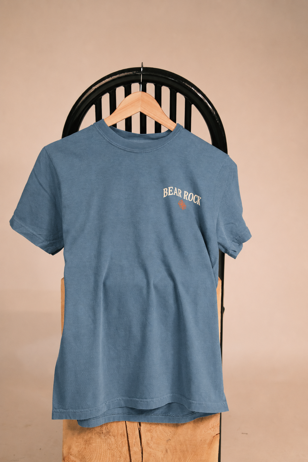 A blue short-sleeve t-shirt with 'BEAR ROCK' printed in white on the left chest, hanging on a wooden hanger from a black chair against a beige background.