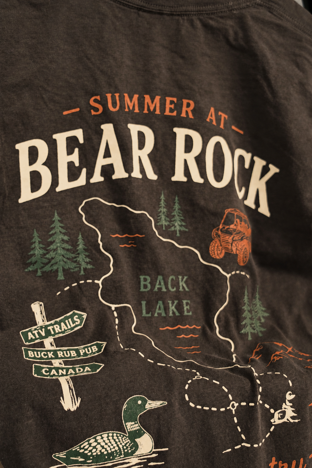 Close-up of a dark charcoal gray t-shirt featuring a 'Summer at Bear Rock Back Lake' graphic print with a map, trees, ATV, duck, and directional signs.