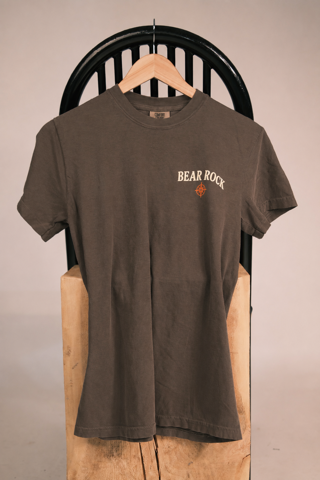 A dark brown short-sleeve t-shirt with 'BEAR ROCK' printed in cream on the left chest, hanging on a wooden hanger from a black metal chair.