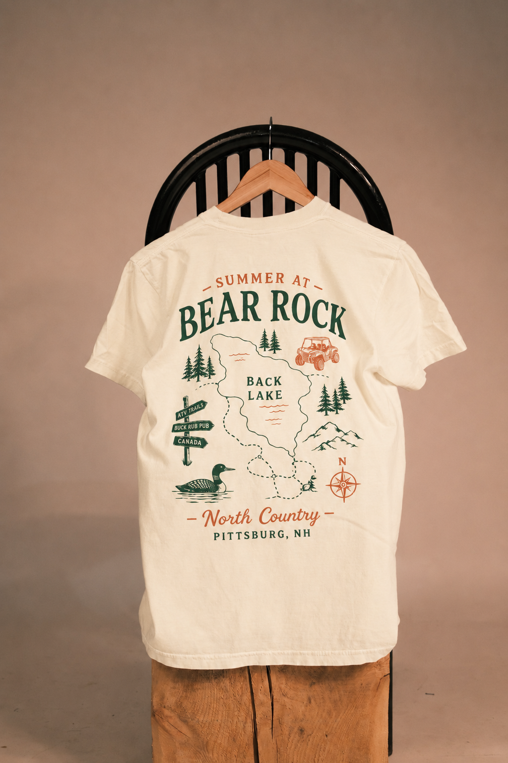 An off-white short-sleeve t-shirt on a wooden hanger, displayed from the back. The shirt features a large graphic print with the text 'SUMMER AT BEAR ROCK - BACK LAKE - North Country - PITTSBURG, NH' in dark green and rust orange, accompanied by illustrations of pine trees, a duck, a car, and a compass rose.
