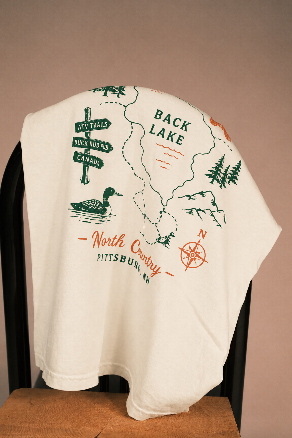 An off-white short-sleeve T-shirt draped over a black chair, featuring a green and orange back graphic with a map of Back Lake, a duck, trees, and directional signs.
