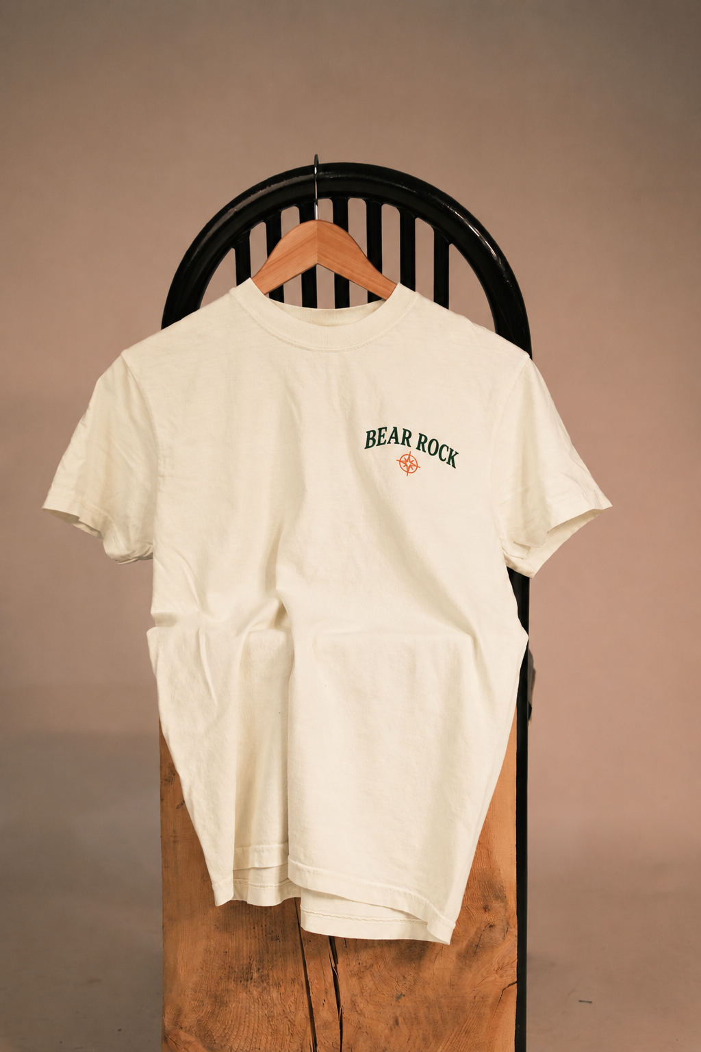 An off-white short-sleeve t-shirt with a green 'BEAR ROCK' graphic on the left chest, hanging on a wooden hanger from a black chair against a warm beige background.