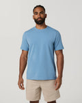 A man wearing a light blue crew-neck t-shirt and beige shorts, standing against a plain light gray studio background.