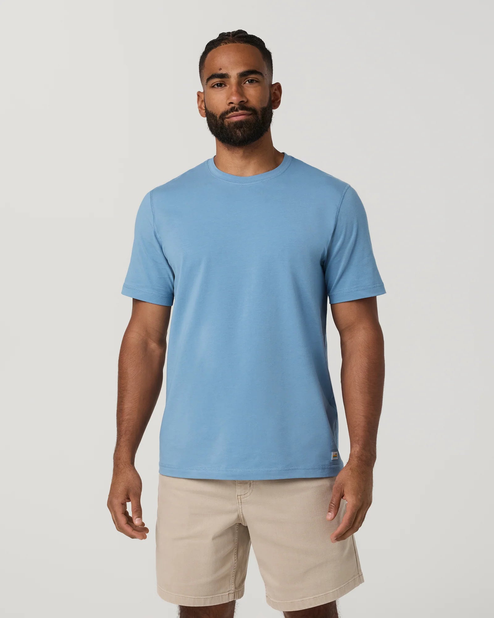 A man wearing a light blue crew-neck t-shirt and beige shorts, standing against a plain light gray studio background.