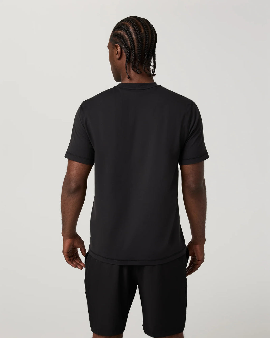 A man viewed from the back wearing a black short-sleeve t-shirt and black shorts against a light gray studio background.