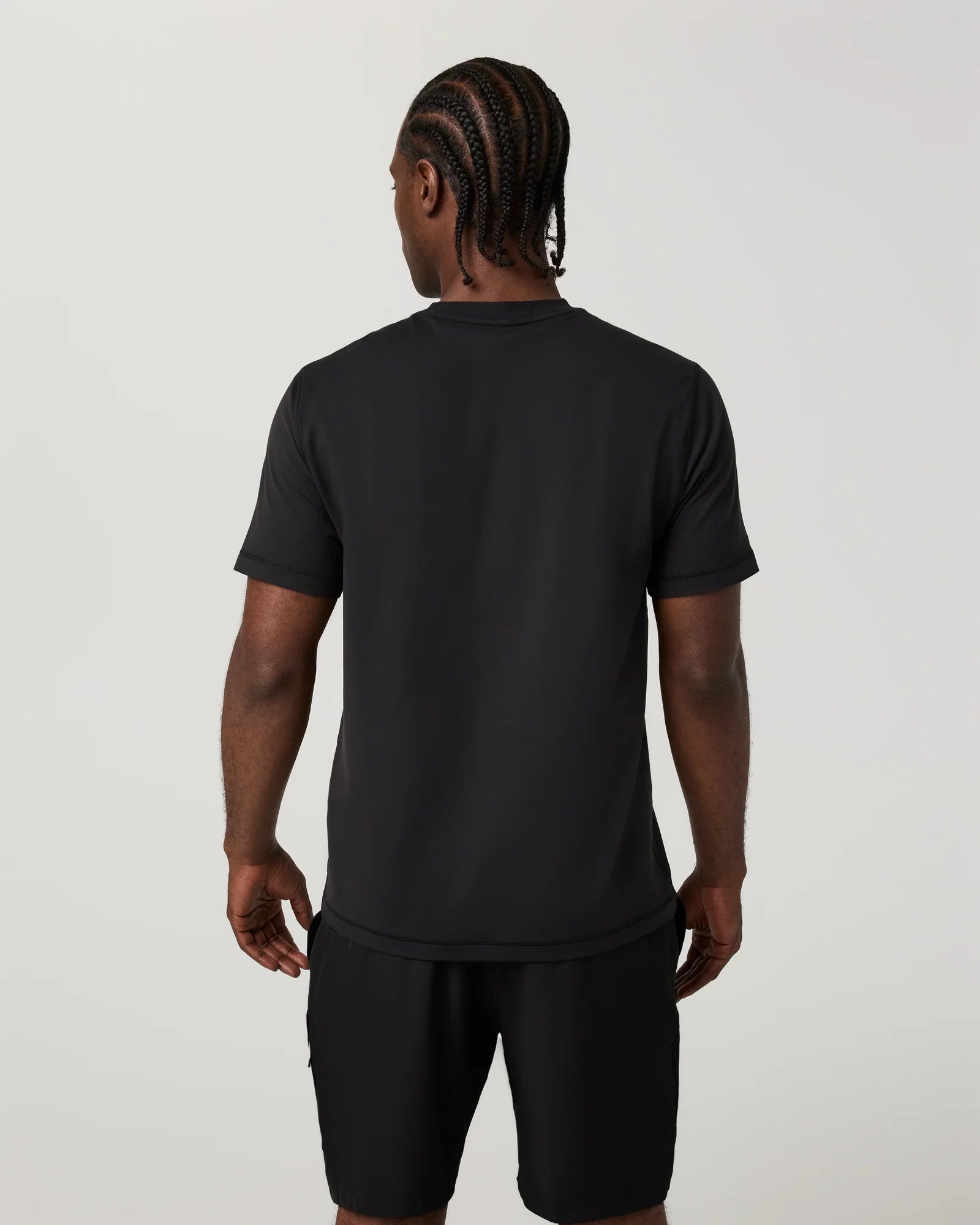 A man viewed from the back wearing a black short-sleeve t-shirt and black shorts against a light gray studio background.