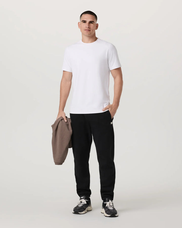 Man wearing a white t-shirt, black pants, and sneakers holding a beige jacket on a light gray background