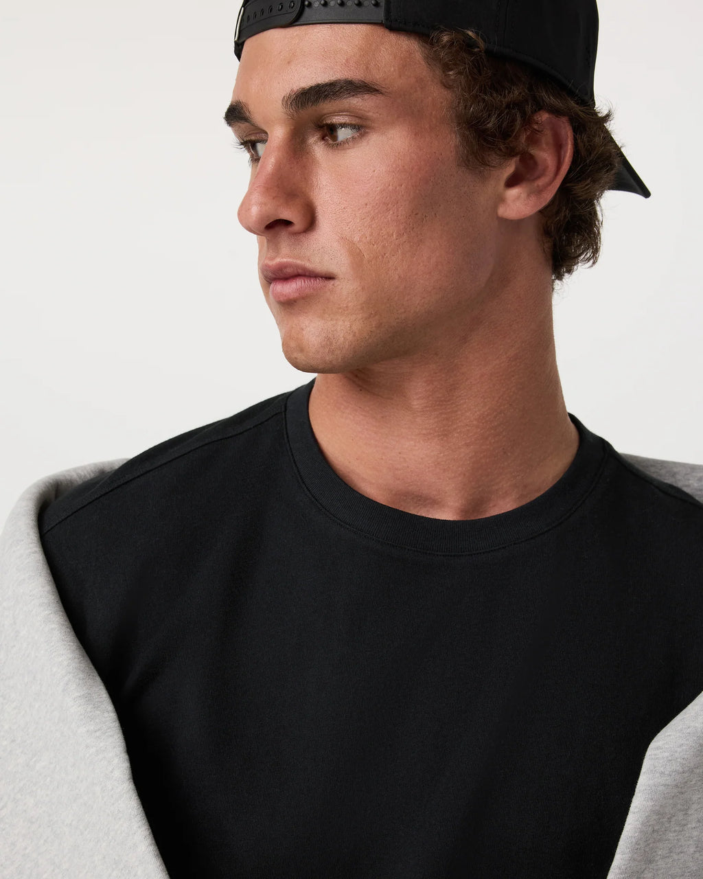 A close-up side profile of a man wearing a black crewneck sweatshirt with light gray sleeves, paired with a black baseball cap, against a plain light gray background.