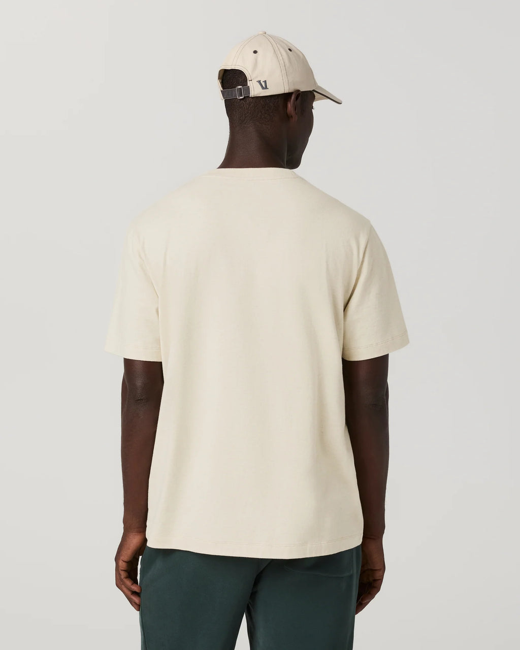 A man shown from the back wearing a cream-colored short-sleeve t-shirt, a matching cream baseball cap, and dark green pants, set against a plain light gray studio background.