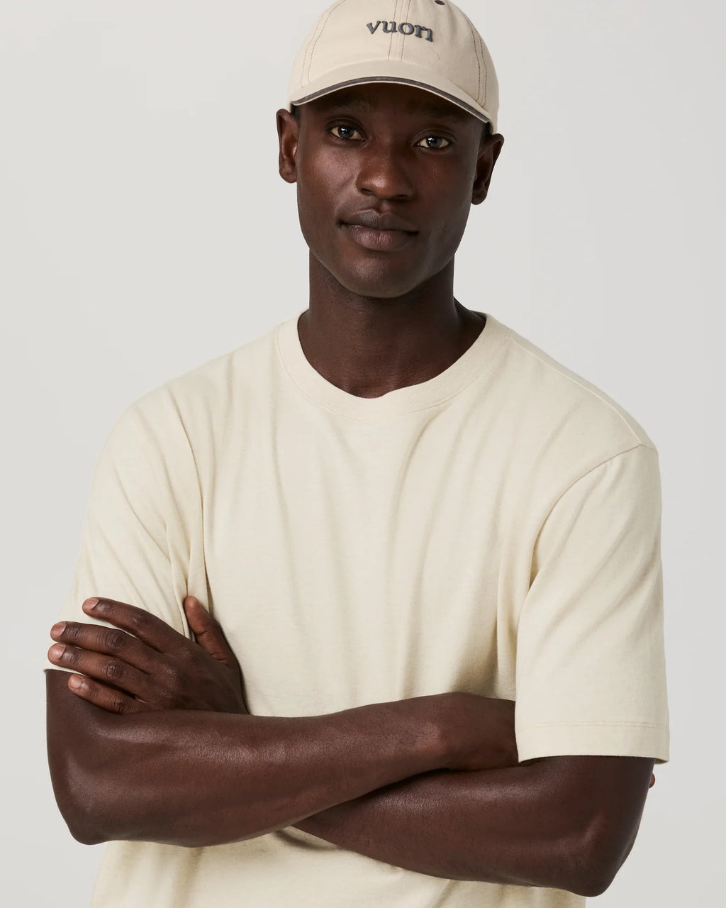 A man wearing a cream-colored short-sleeve t-shirt and a matching cream-colored Vuori baseball cap, standing with his arms crossed against a light gray studio background.