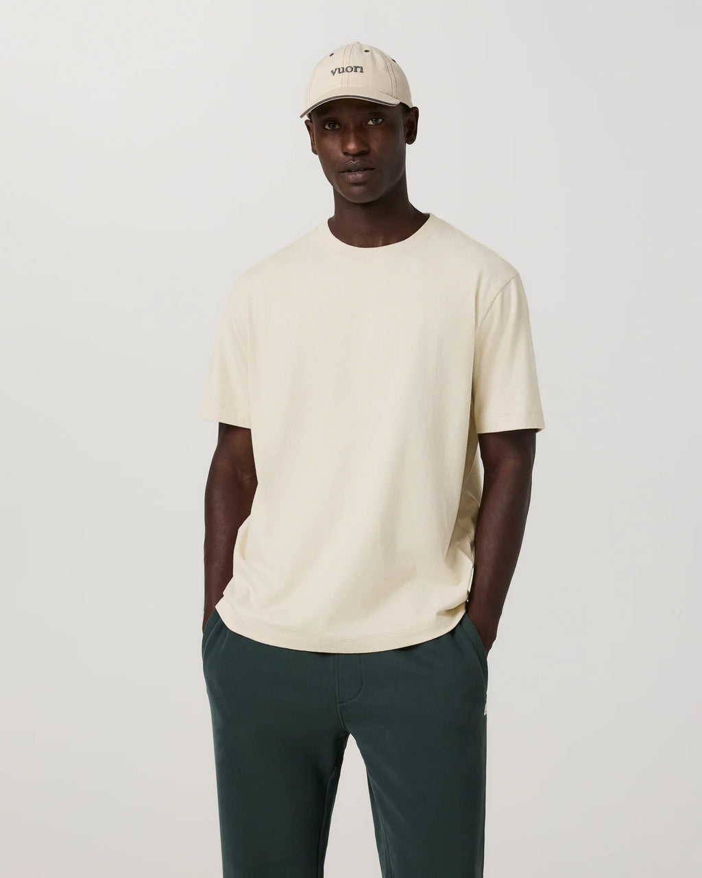 A man modeling a cream-colored short-sleeve crewneck t-shirt, paired with dark green trousers and a matching cream cap, against a plain light gray background.
