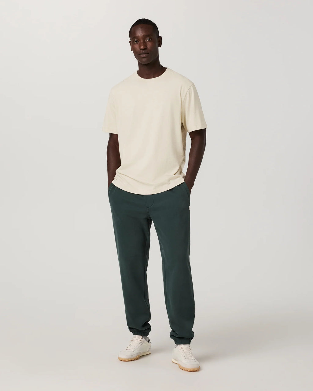 A man modeling a cream short-sleeve crewneck t-shirt and dark green jogger pants with white sneakers, standing against a light gray studio background.