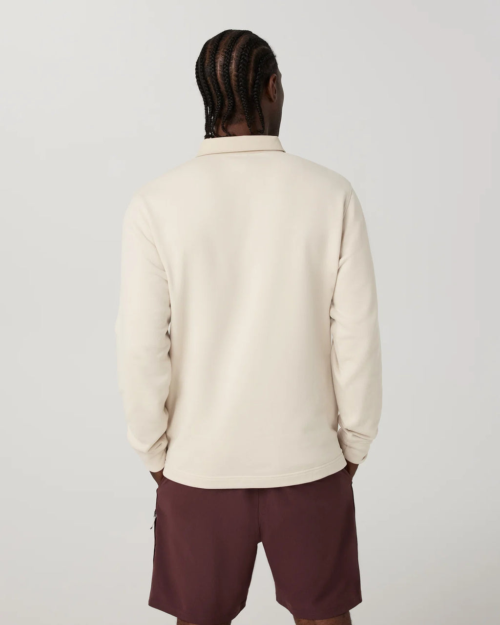 A man viewed from behind wearing a cream-colored long-sleeve collared shirt and dark maroon shorts, standing against a light gray studio background.