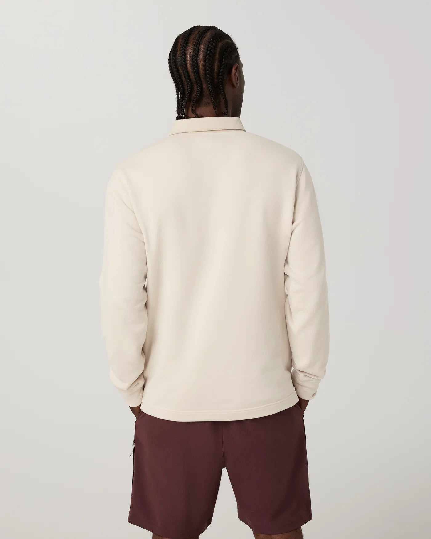 A man viewed from behind wearing a cream-colored long-sleeve collared shirt and dark maroon shorts, standing against a light gray studio background.