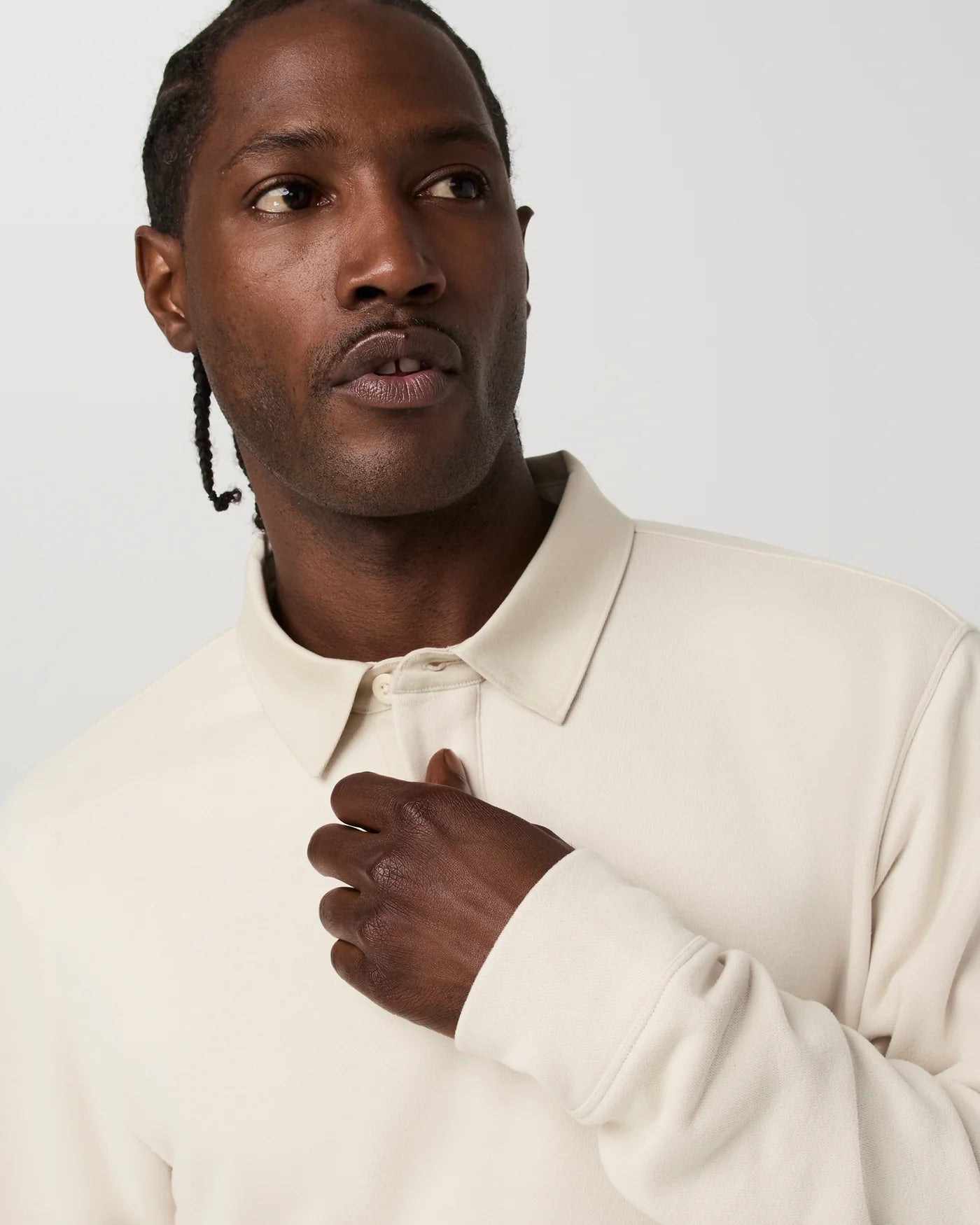 A close-up shot of a man wearing a cream-colored long-sleeve polo shirt, looking off-camera with his hand resting on his chest against a light gray background.