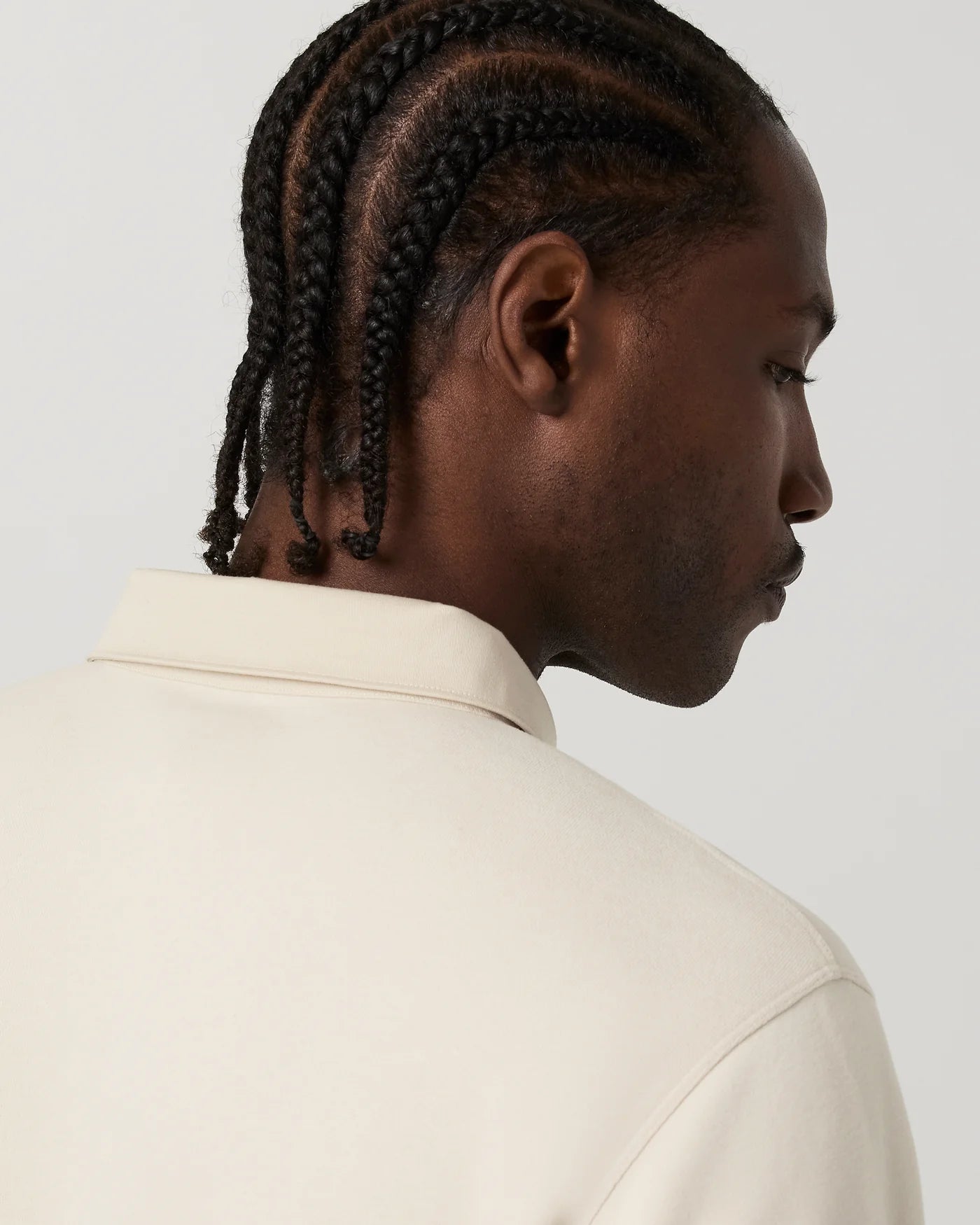 A close-up side profile of a man with dark braided hair wearing a cream-colored collared shirt against a light gray background.