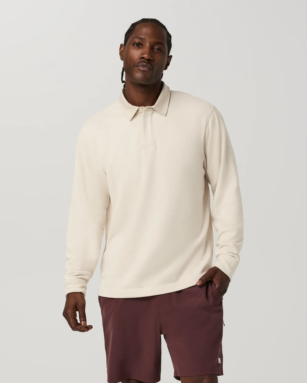 A man wearing a cream long-sleeve polo shirt and burgundy shorts, standing against a light gray studio background.