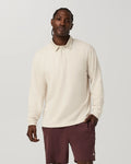 A man wearing a cream long-sleeve polo shirt and burgundy shorts, standing against a light gray studio background.