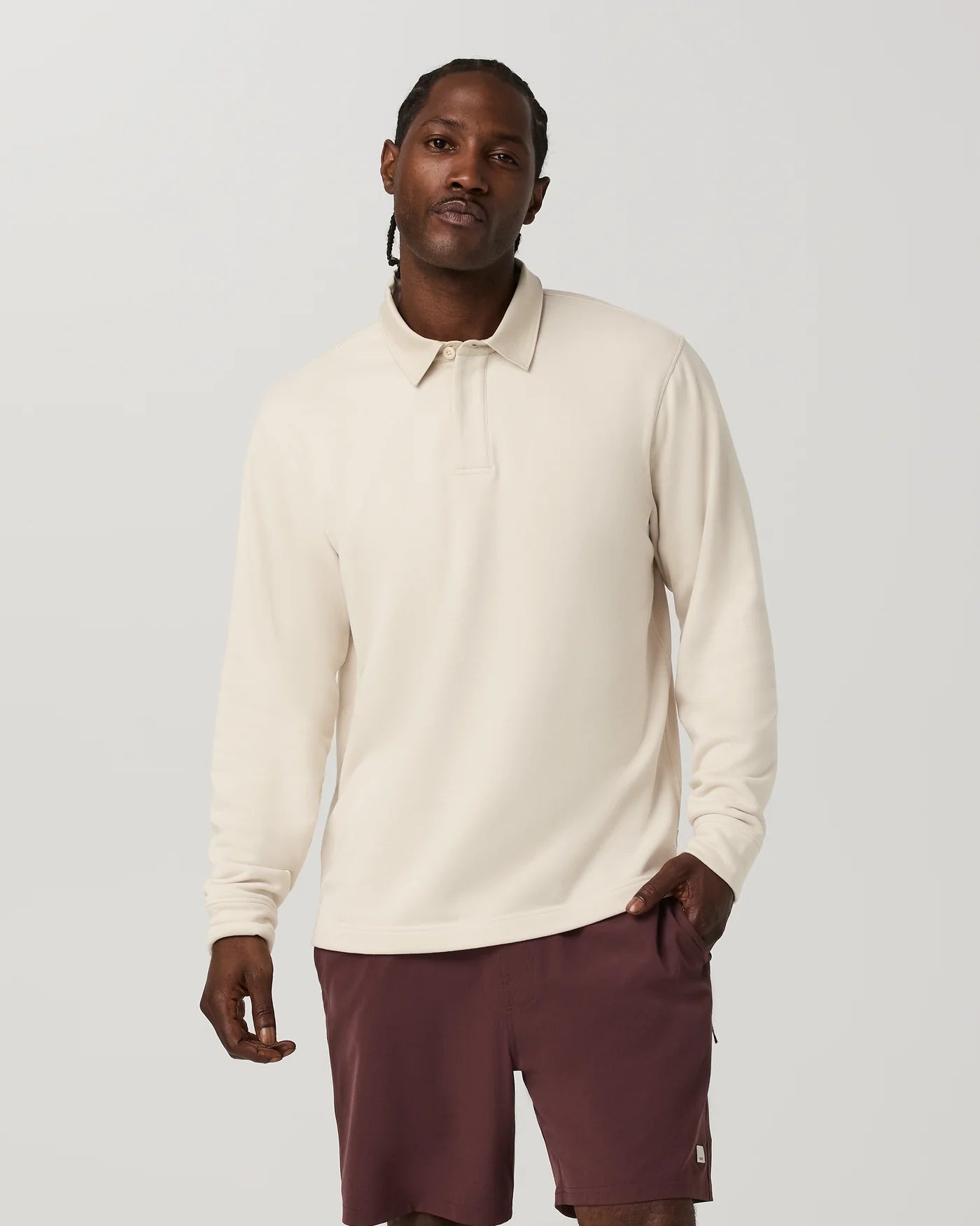 A man wearing a cream long-sleeve polo shirt and burgundy shorts, standing against a light gray studio background.