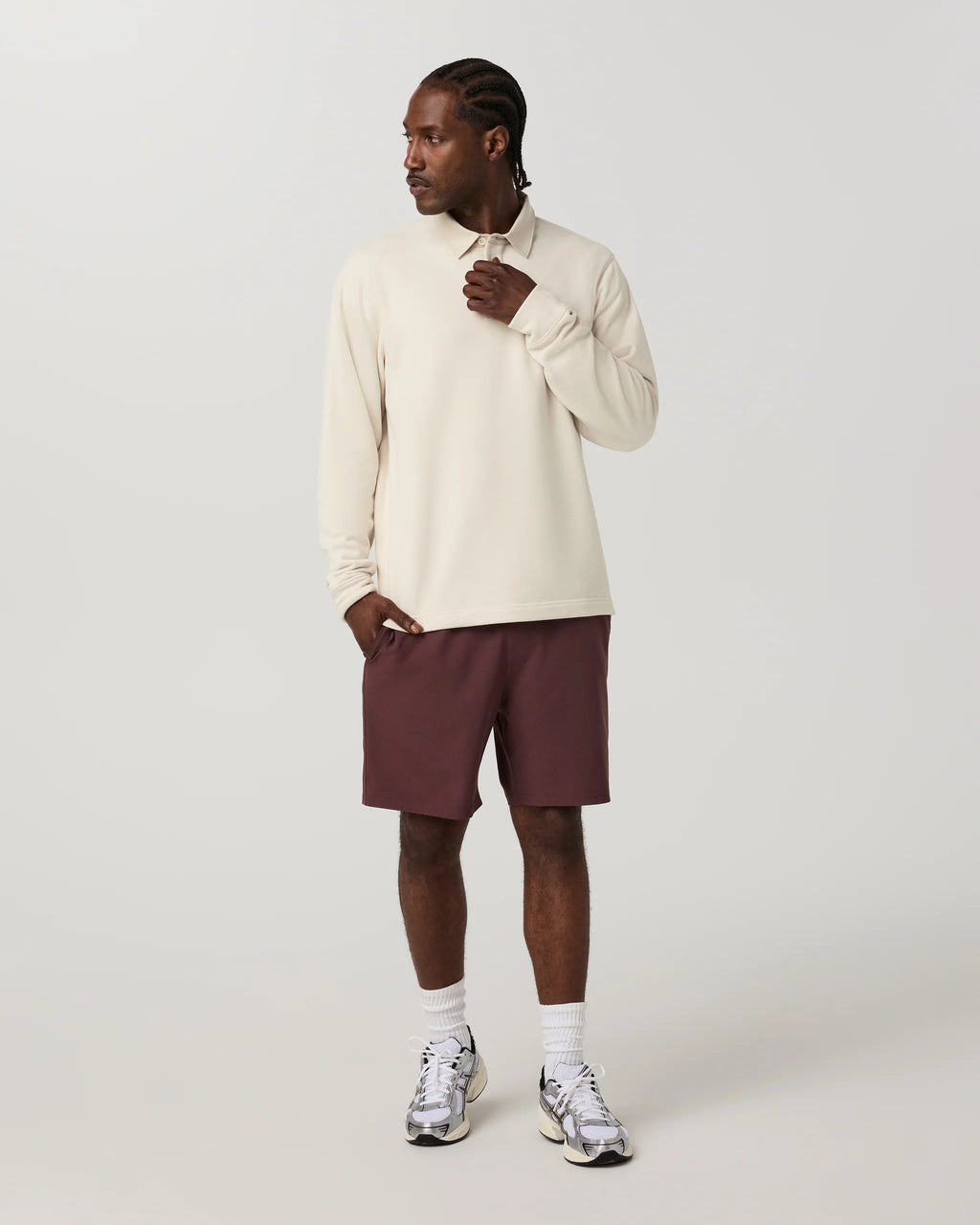 A man modeling a cream long-sleeve collared shirt with dark burgundy athletic shorts, white socks, and silver sneakers against a light gray studio background.