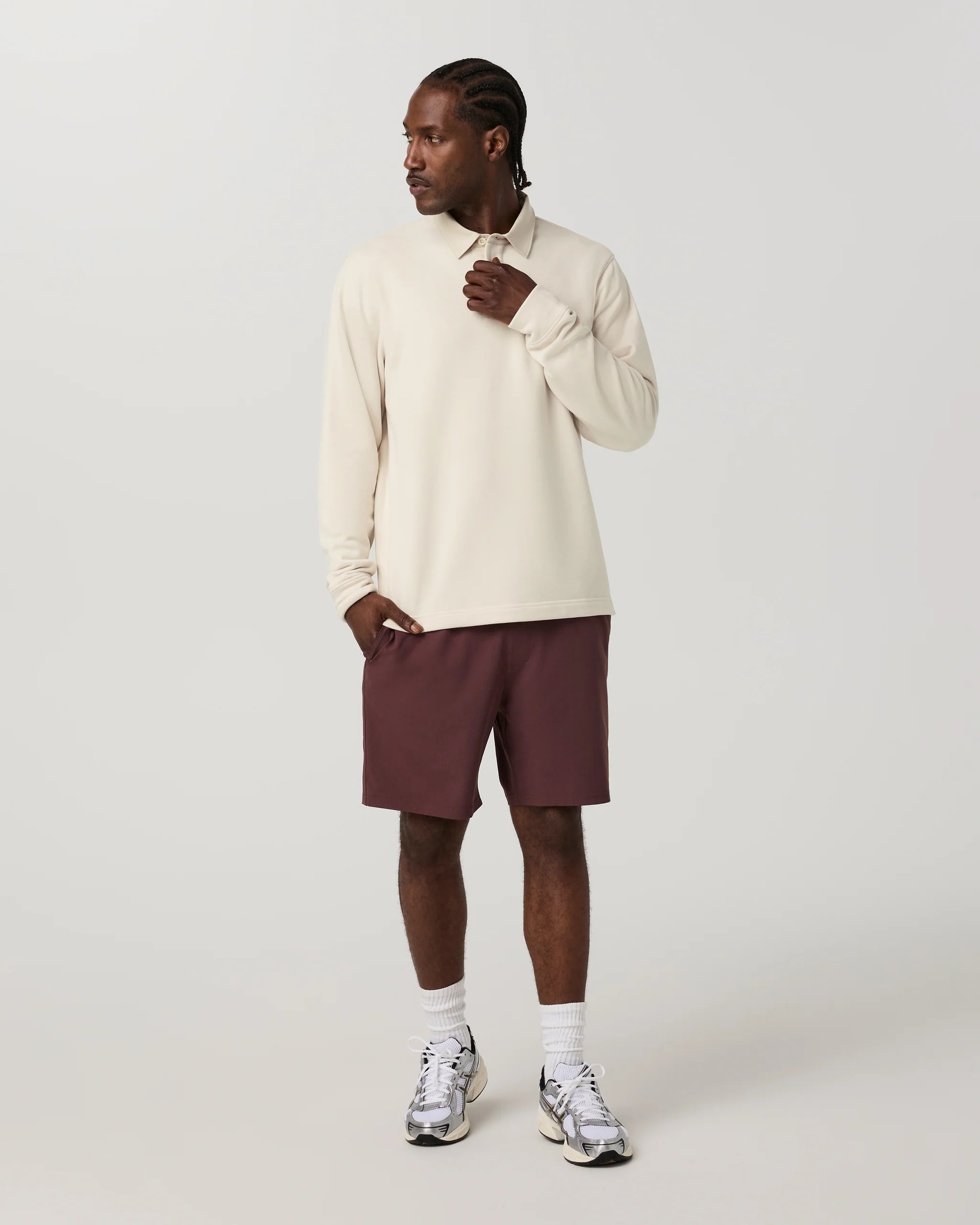 A man modeling a cream long-sleeve collared shirt with dark burgundy athletic shorts, white socks, and silver sneakers against a light gray studio background.