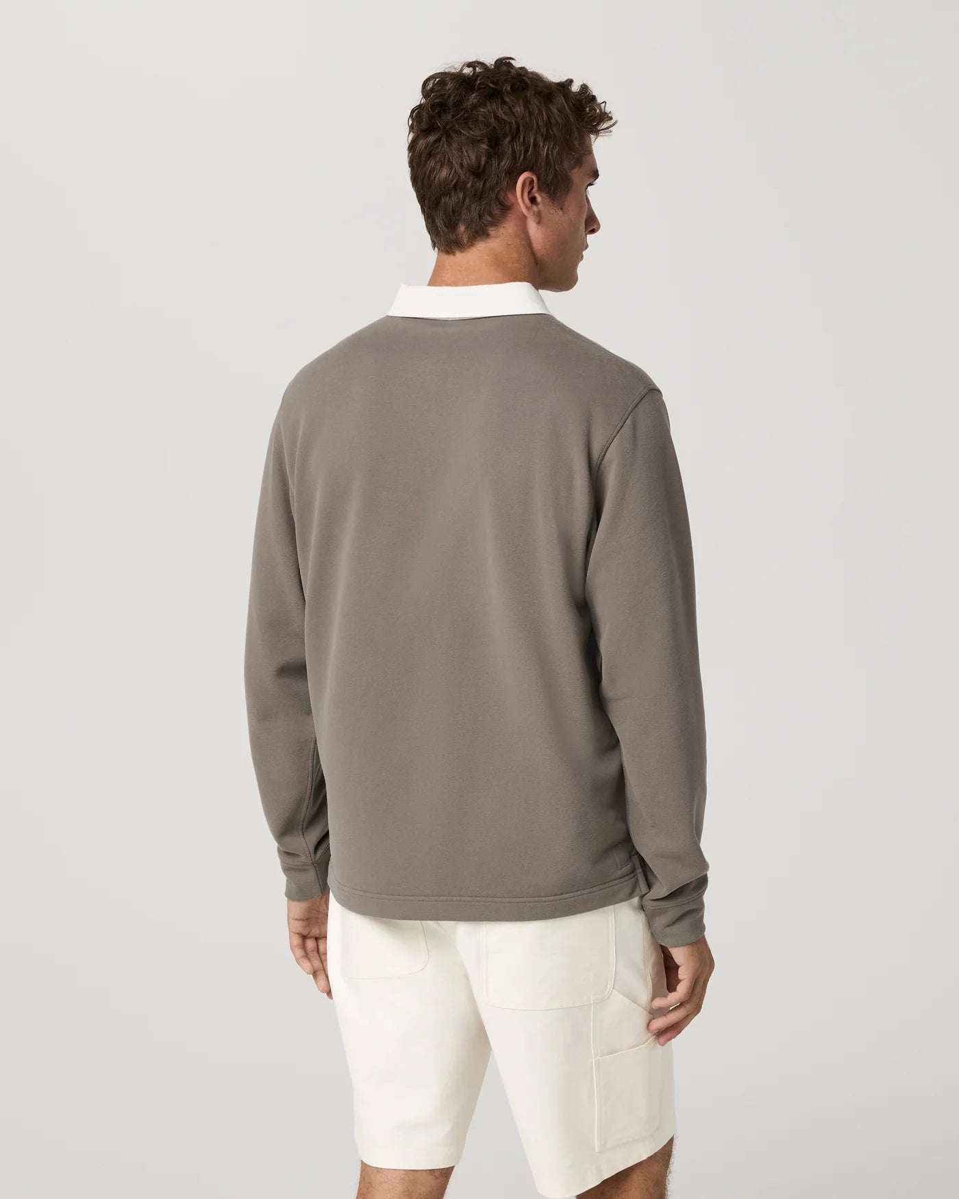 A man shown from the back wearing a taupe-brown long-sleeve crewneck sweatshirt layered over a white collared shirt, paired with off-white shorts, against a light gray studio background.
